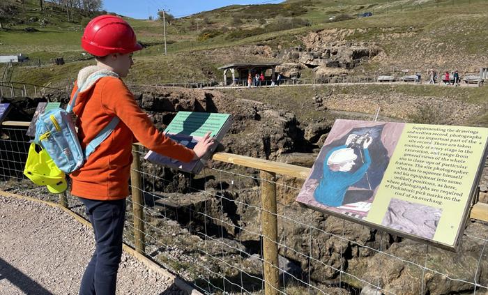 A child is standing reading an information board above a chasm in the rock below.  In the distance people are walking on the path on the other side an